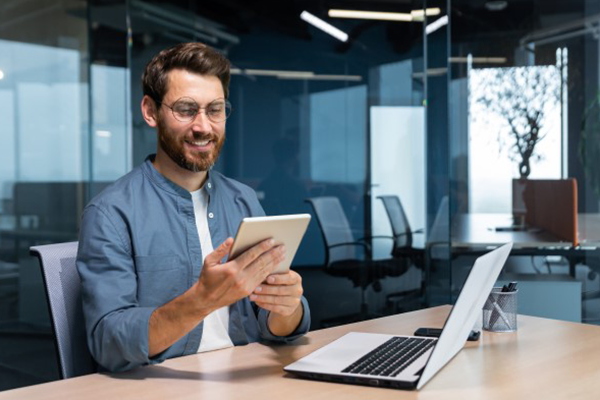 Pan spoglada na ekran tableta, siedząc przy biurku w biurze. Na stole leży otwarty laptop.