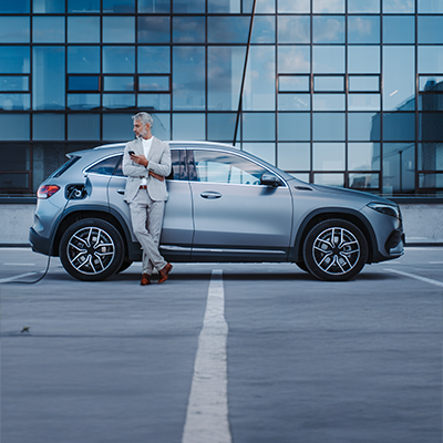 Mężczyzna podłączający przewód ładowania do srebrnego samochodu elektrycznego przed przeszklonym budynkiem.