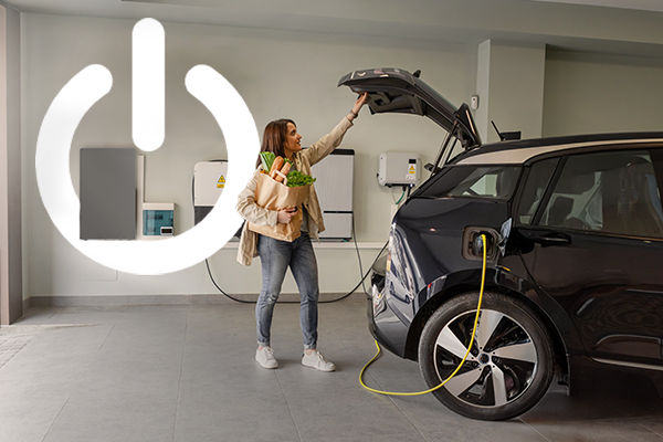kobieta w garażu wyciąga zakupy z samochodu elektrycznego, w tle magazyn energii i stacja ładowania samochodó elektrycznych