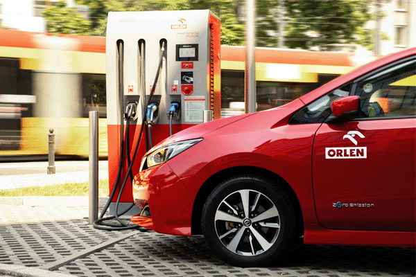 Czerwone auto elektryczne ładuje się przy stacji ORLEN Charge, w tle widać przejeżdżający tramwaj
