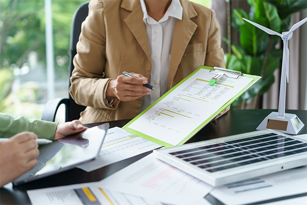 Uścisk dłoni dwóch mężczyzn na tle kobiety trzymającej tablet; jeden z mężczyzn trzyma panel fotowoltaiczny.