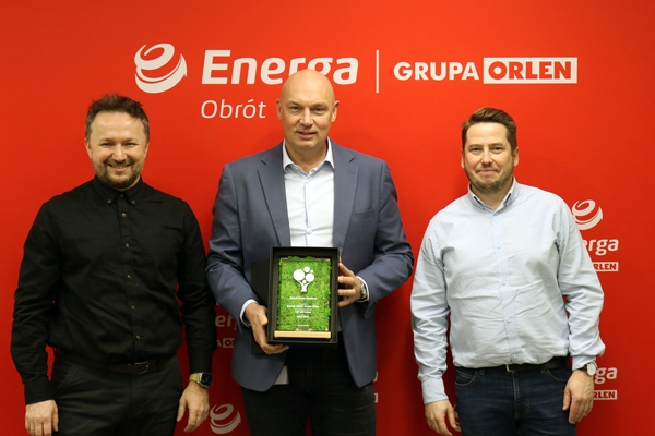 Trzech eleganckich, uśmiechniętych mężczyzn, jeden z nich trzyma certyfikat w ramce. W tle czerwony baner z logo Energa Obrót, Grupa ORLEN.