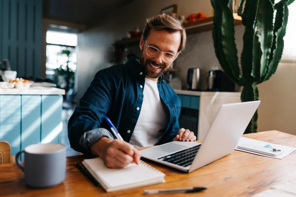 Osoba pracuje w domu przy drewnianym stole, jednocześnie korzystając z laptopa i zapisując notatki w zeszycie. Obok stoi kubek, a w tle widać domowe wnętrze z kuchenną zabudową, co tworzy spokojną atmosferę