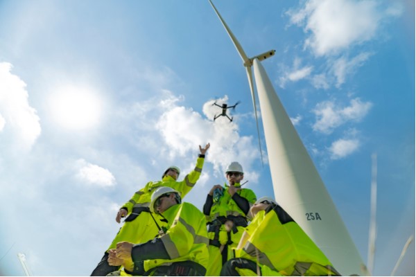 Grupa czterech techników w jaskrawych kamizelkach i kaskach stoi pod wysoką turbiną wiatrową, obserwując lecącego drona na tle błękitnego nieba.