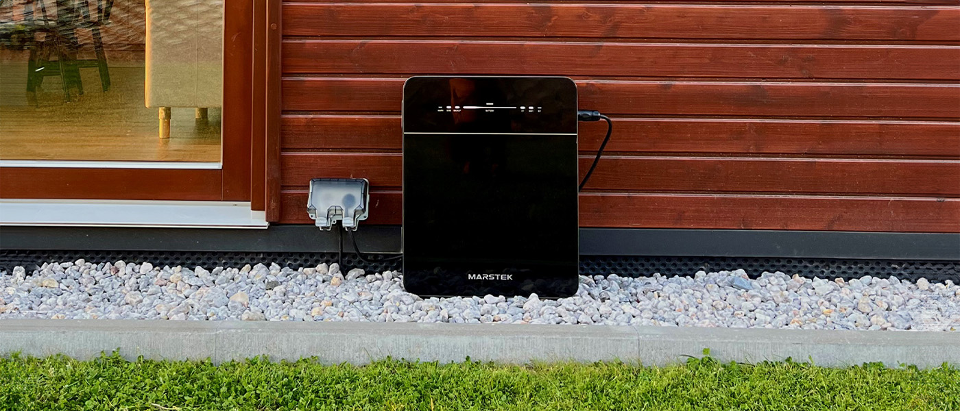 Magazyn energii Marstek Venus stoi przed domem na kamyczkach, w tle drewniana elewacja oraz wnętrze domu widoczne przez okno balkonowe.