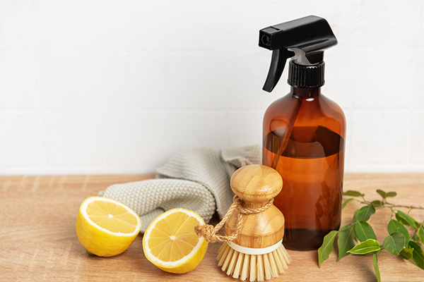 Butelka z atomizerem stoi na blacie, obok przekrojone połówki cytryny, ściereczka, szczotka do czyszczenia oraz zielone liście