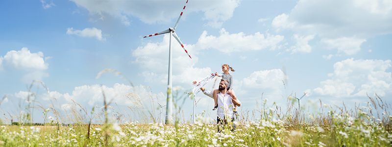 Ojciec z córką stoją na łące. W tle turbiny wiatrowe.