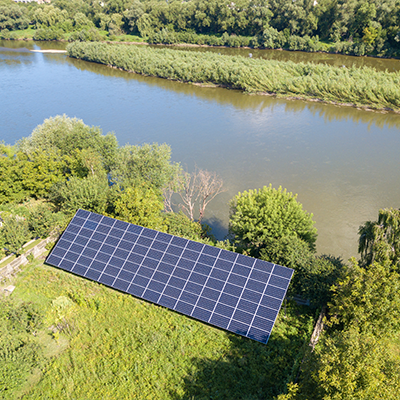 Widok z góry na panele fotowoltaiczne umiejscowione nad brzegiem rzeki w otoczeniu lasu.